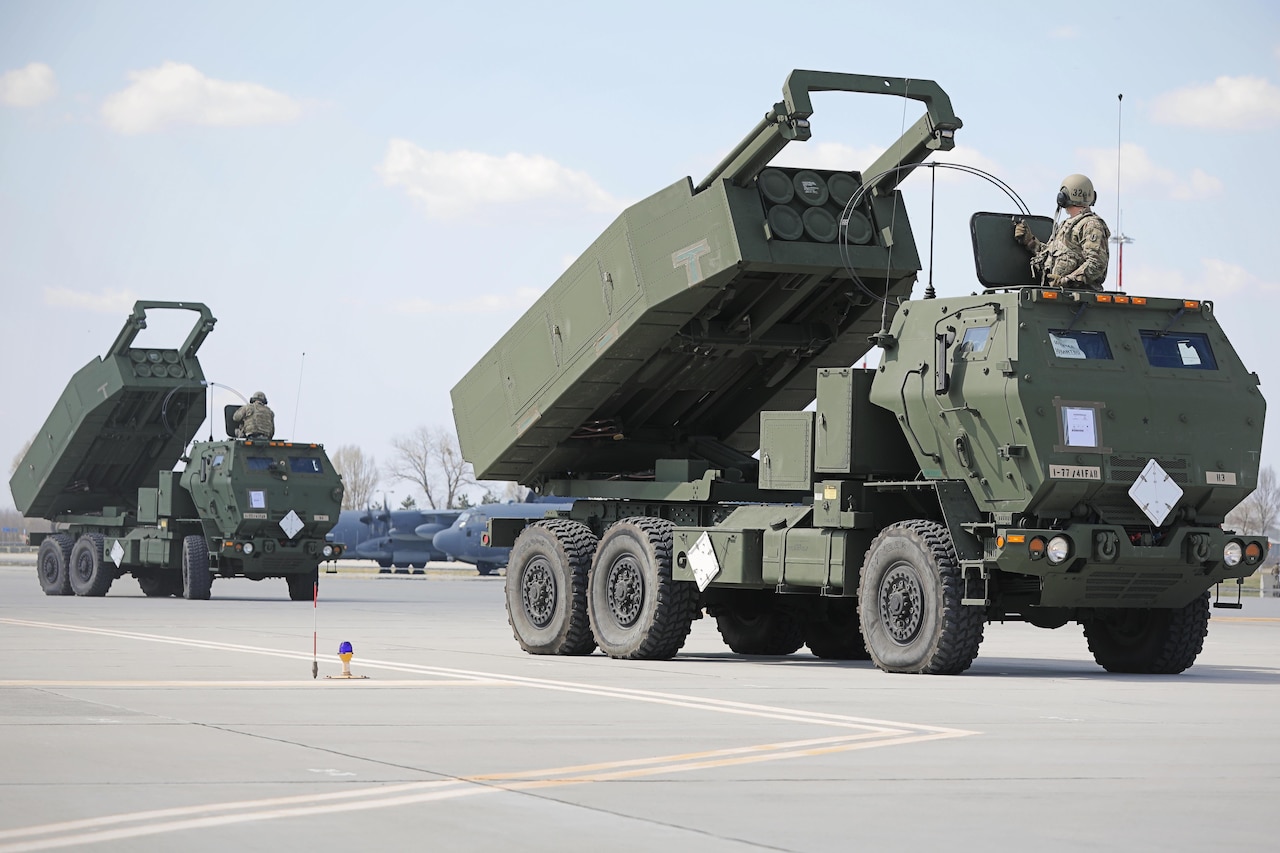 Two trucks containing artillery are parked on the pavement.