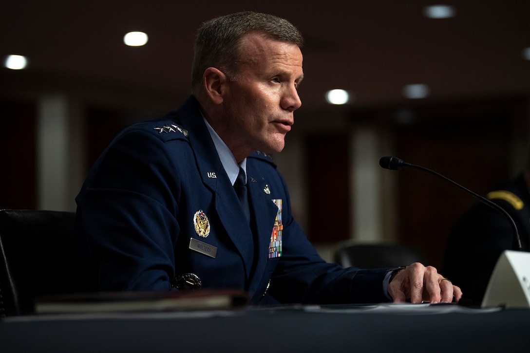 A man dressed in a military uniform sits at a table and speaks into a microphone.