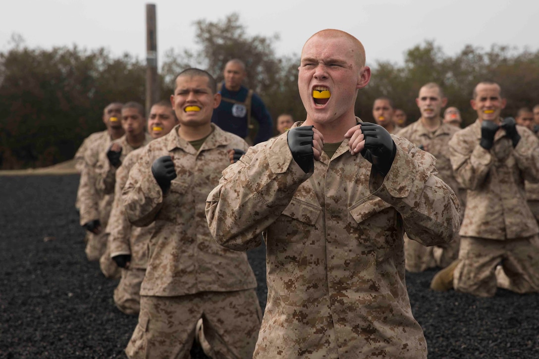 A group of Marine Corps recruits kneel on the ground while raising their hands.