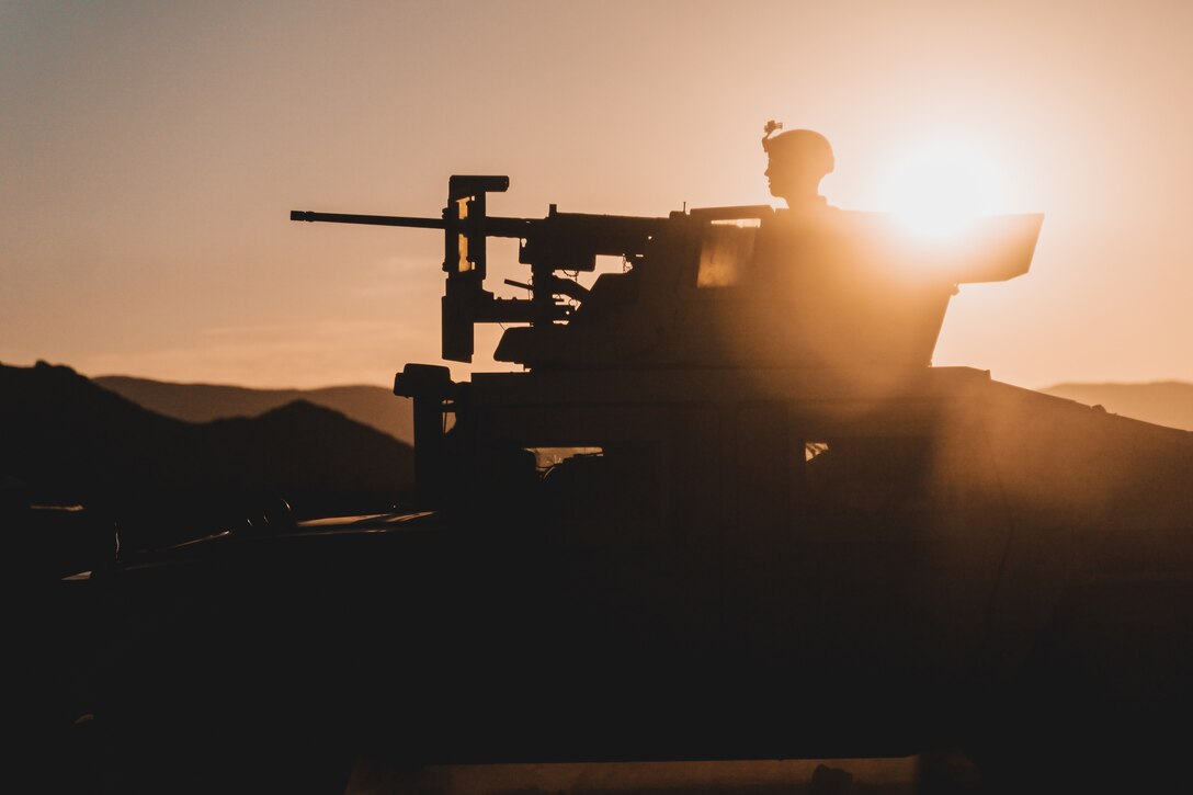 A Marine sits in a tank under a sunlit sky as shown in silhouette.