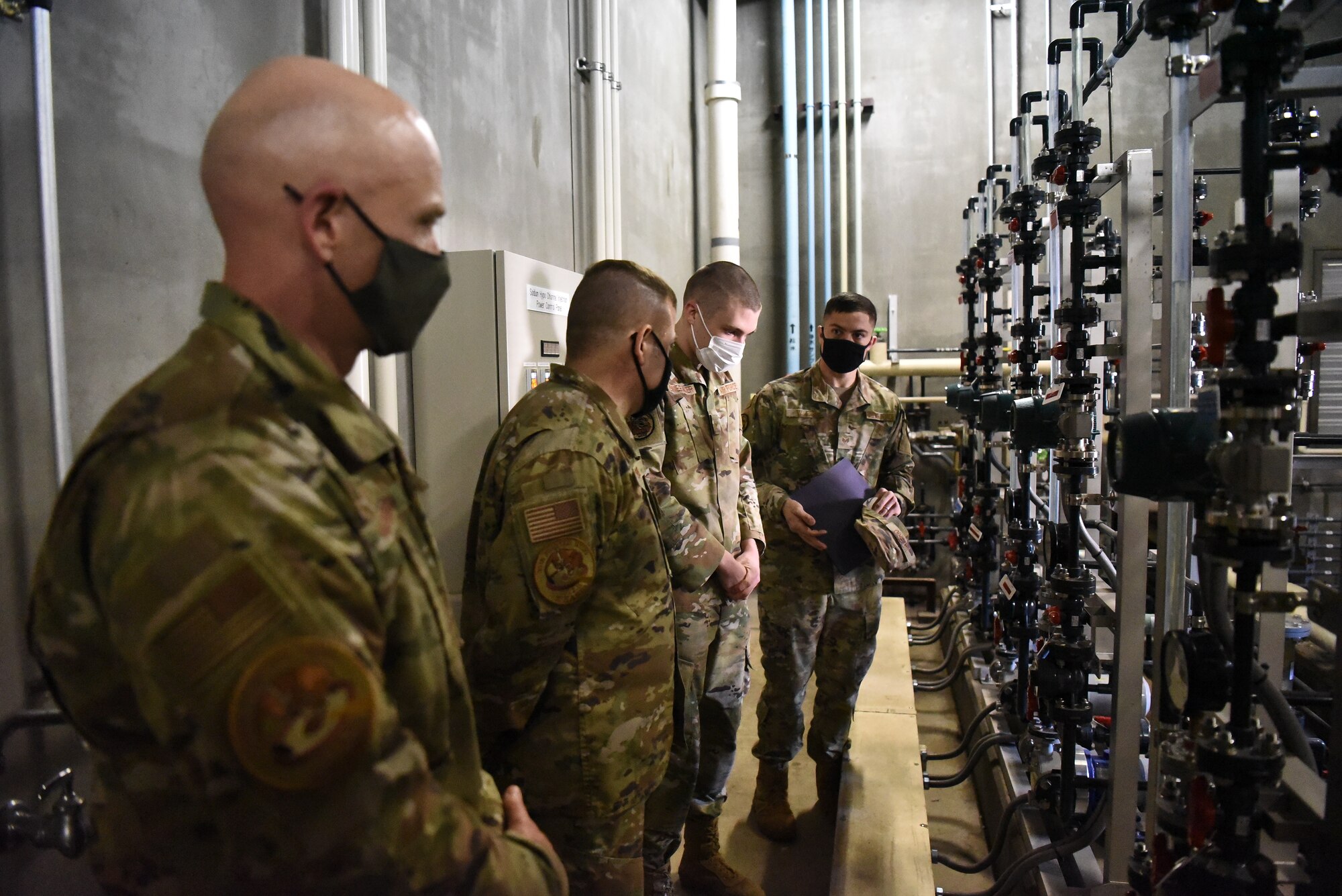 Four men in uniform stand looking at large pumps and equipment on the wall.