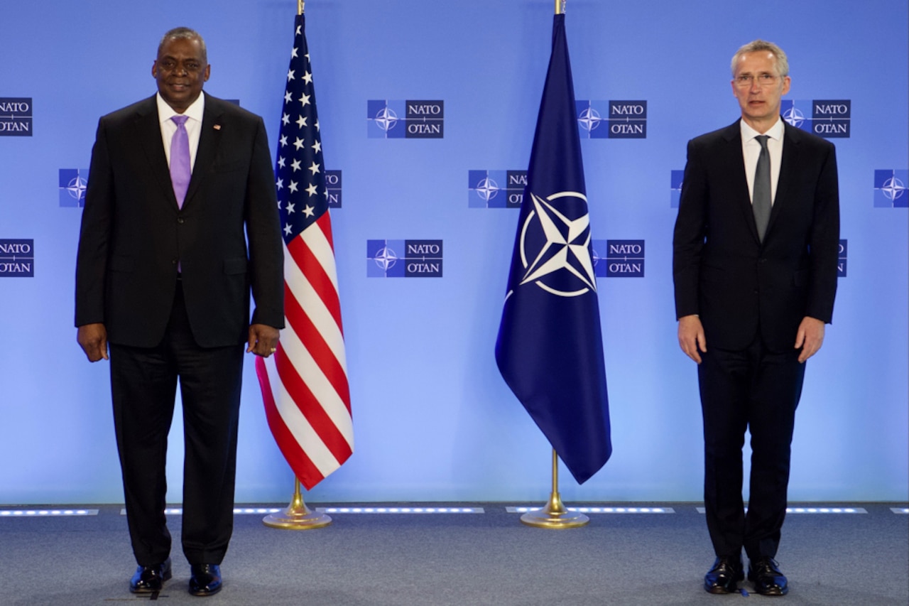 Two men stand beside each other next to U.S. and NATO flags.