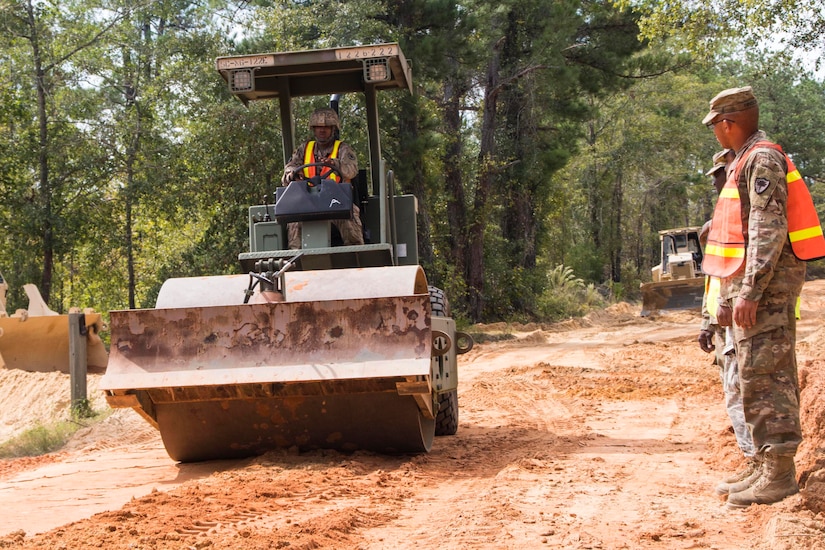 A man in a uniform operates a rolling compactor as it travels down a dirt road. Two other men in uniform stand to the side of the road.
