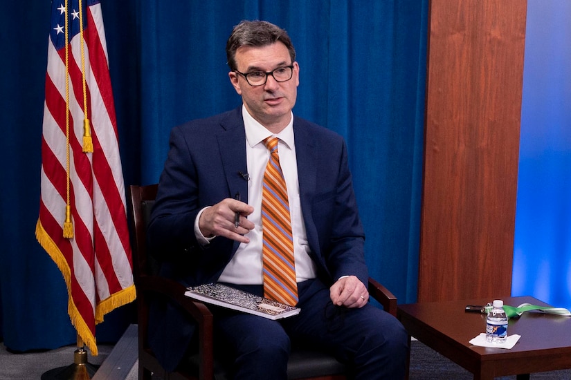 A man seated in a chair speaks. Behind him is an American flag.