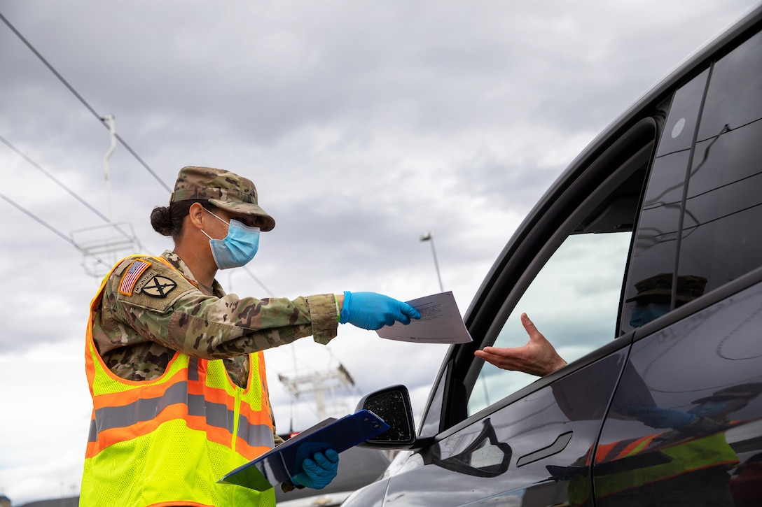 A soldier wearing a face mask and gloves hands a flyer to someone in a vehicle.