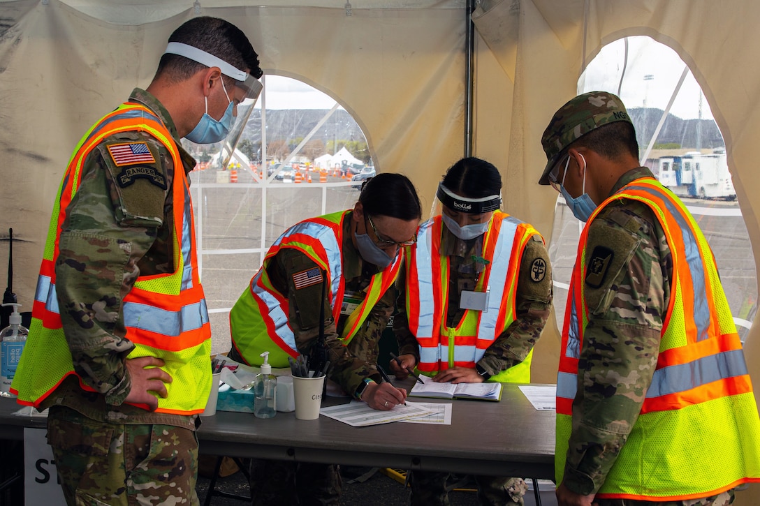 One soldier leans over a table to write on a piece of paper as another reads a ledger and two others watch. All are wearing face masks and gloves.