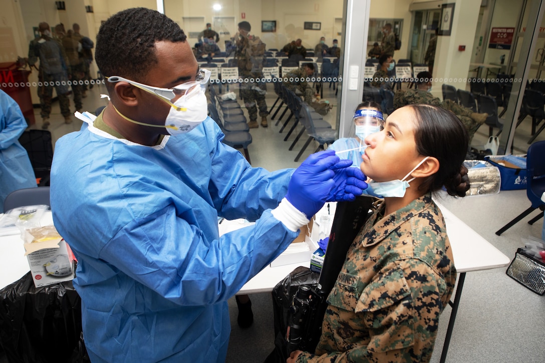 A male sailor wearing a face mask, gloves and protective clothing swabs the nasal passage of a female Marine.