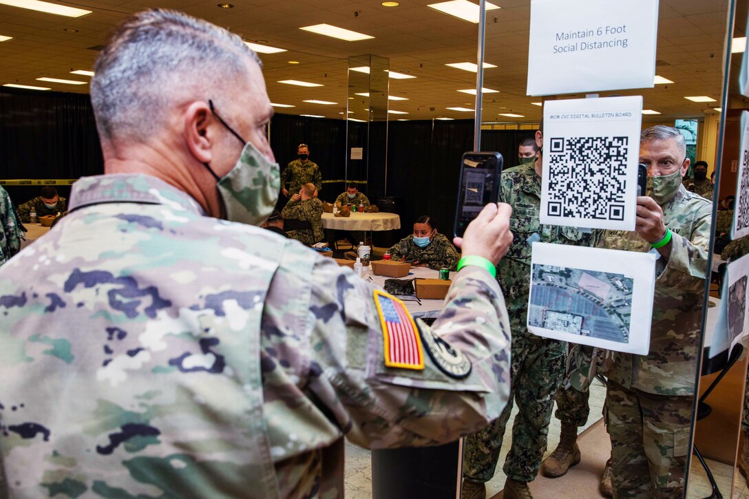 A soldier wearing a face mask holds his phone up to scan a barcode taped to a glass panel in a large room. In the background, soldiers sit at large round tables.