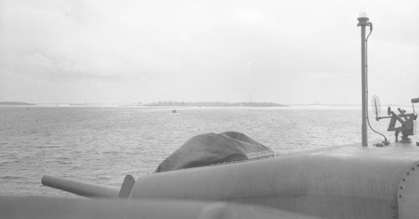 A ship with a large gun sits off the shore of an island.