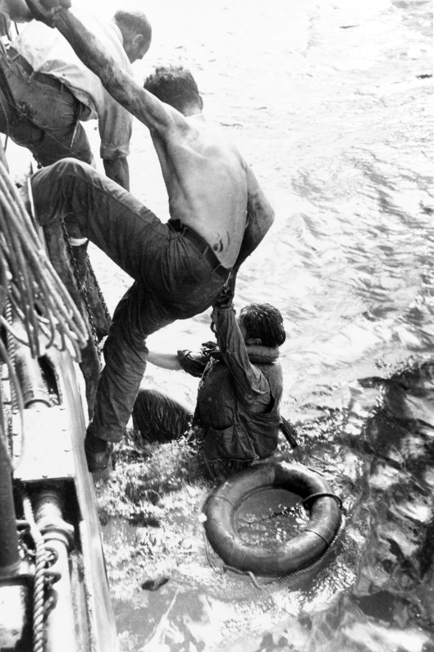 Two men hang onto the side of a ship as they work to pull a third man from the ocean.