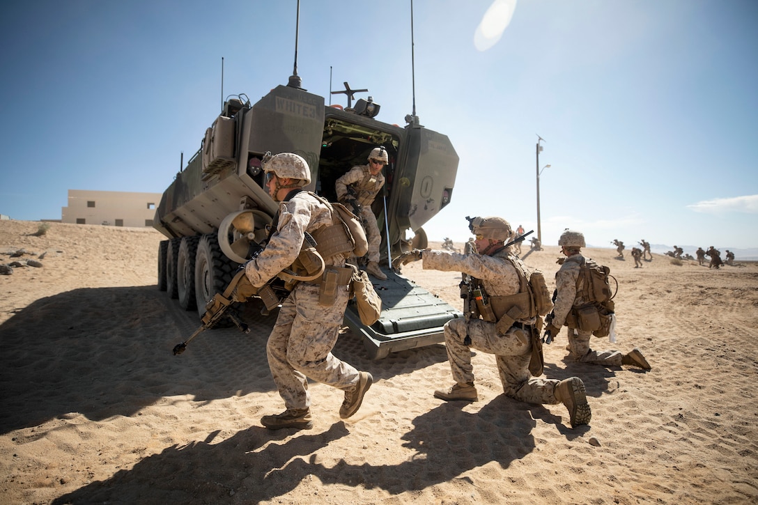 Marines run on sand near a parked amphibious vehicle.