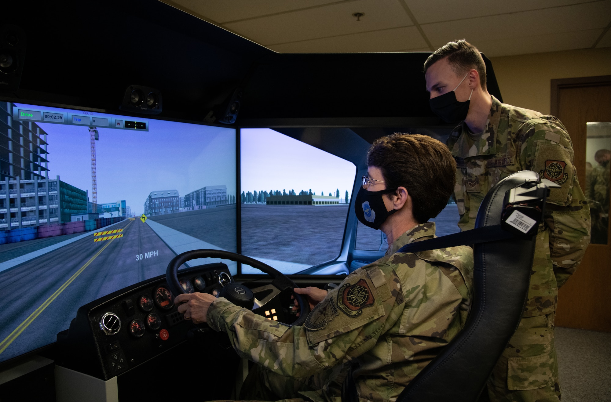 Woman sits in driving simulator while man looks over her shoulder.