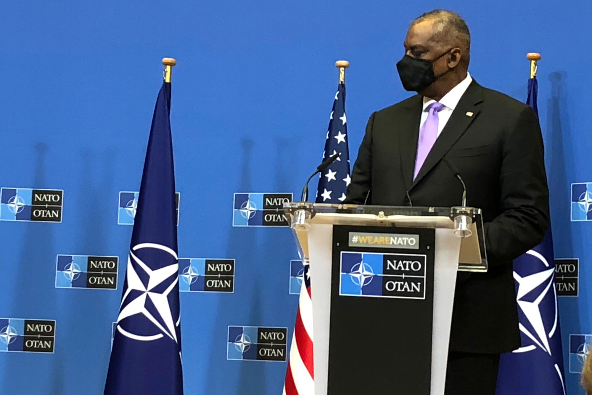 A man in business attire stands at a lectern with microphones and U.S. and NATO flags behind him.