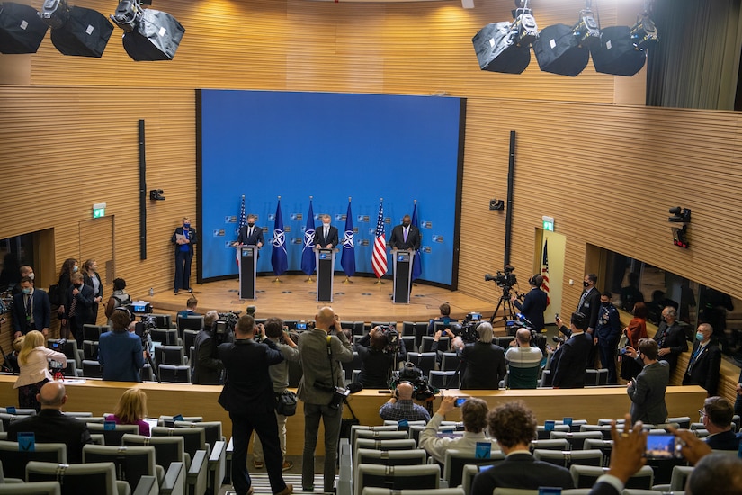 Three men stand on a stage in an auditorium with people in seats in front of them.