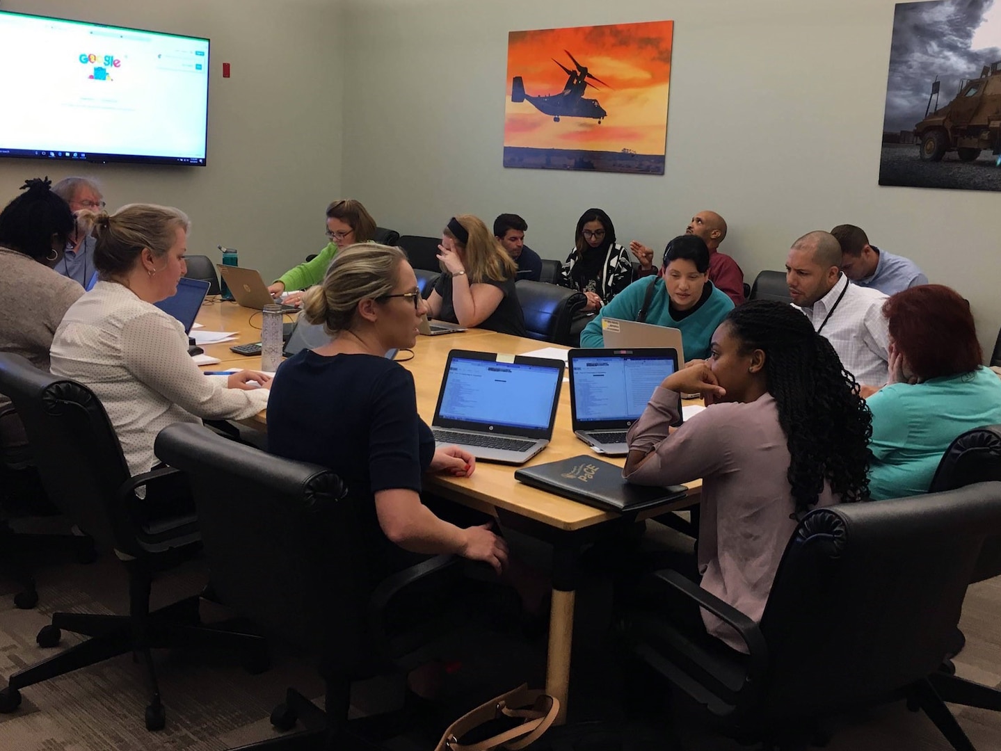 Several professionally dressed people sit around a large conference table with laptops seemingly discussing something.