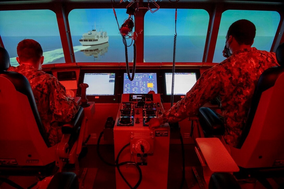 Two sailors illuminated by red light sit inside a simulated ship.