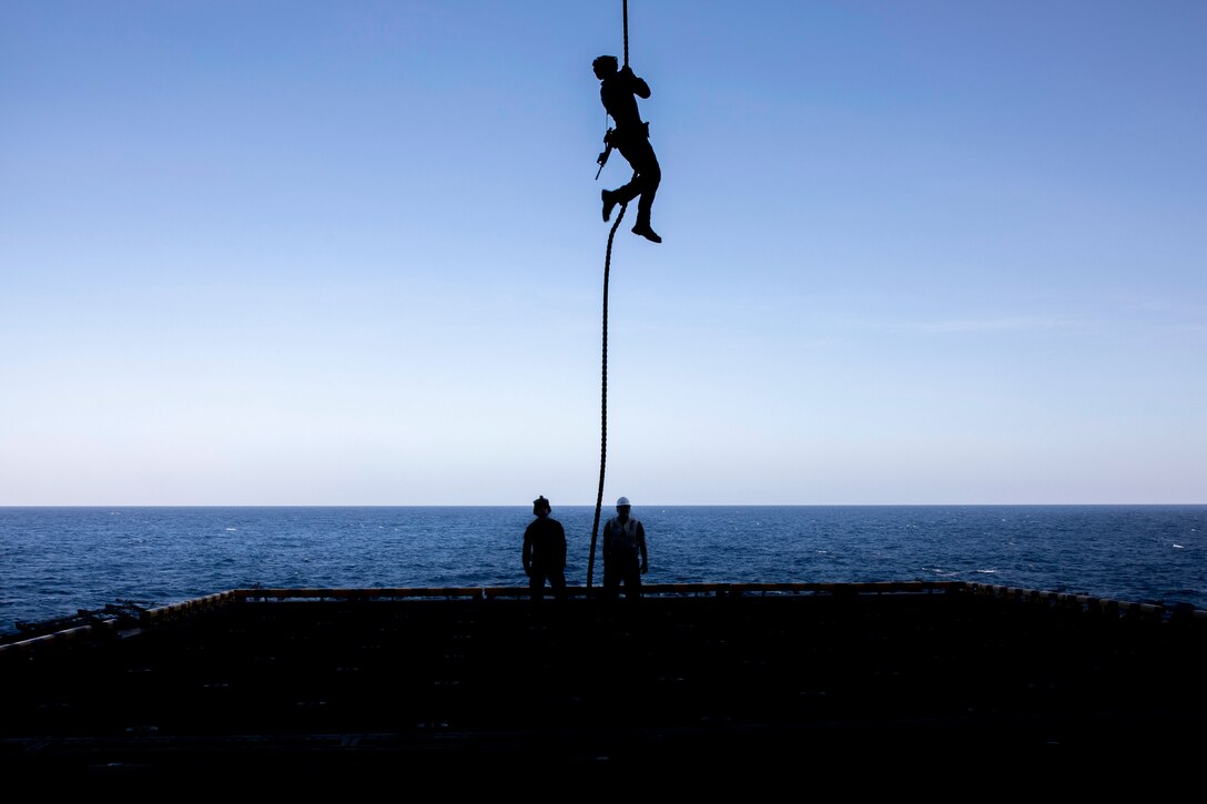 A Marine lowers onto a ship from a rope while fellow Marines watch as shown in silhouette.