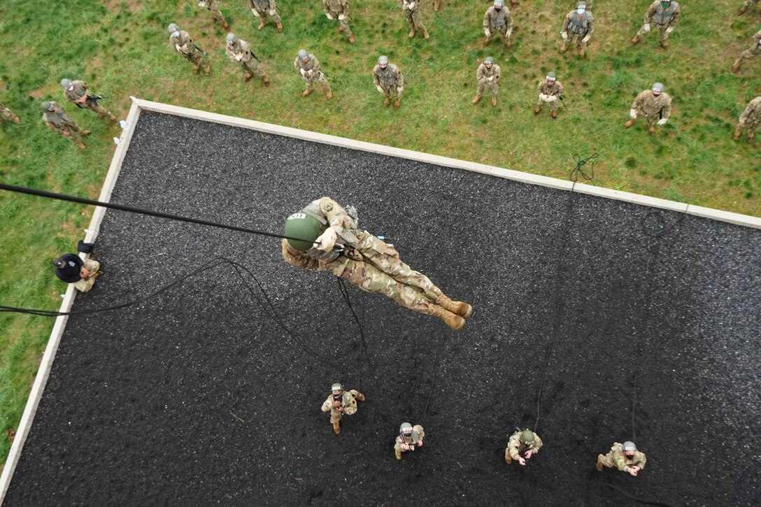 A soldier rappels down on a rope as others watch from below.