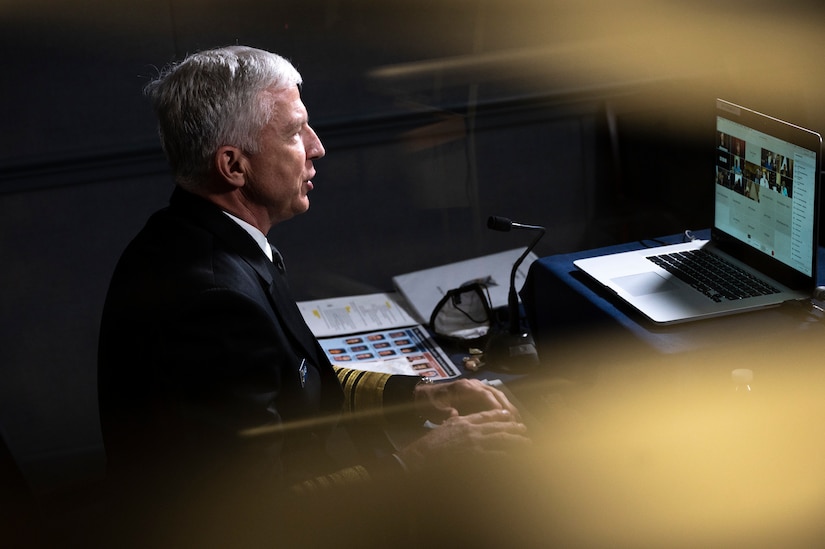 A man in a military uniform interacts with a congressional hearing through his laptop.