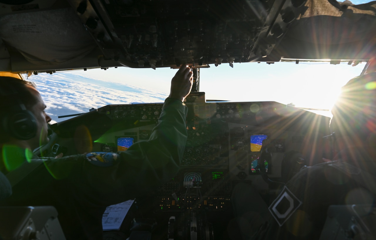 Airmen, shown from behind, sit in the darkened cockpit of an aircraft in flight.