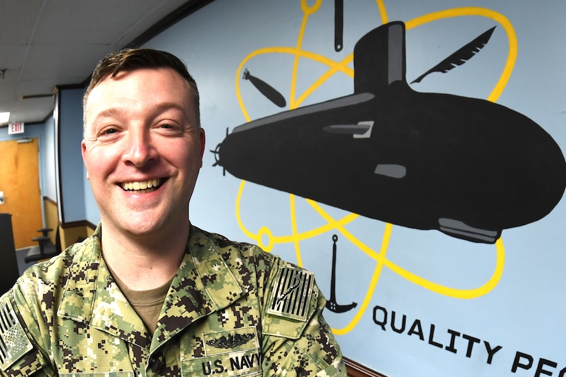 A sailor smiles in front of a painting of a submarine.