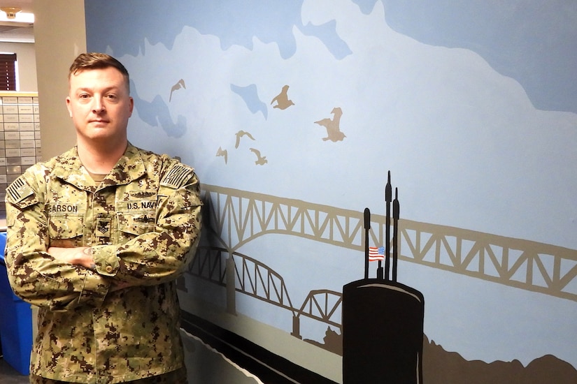 A sailor stands with arms crossed in front of a mural.