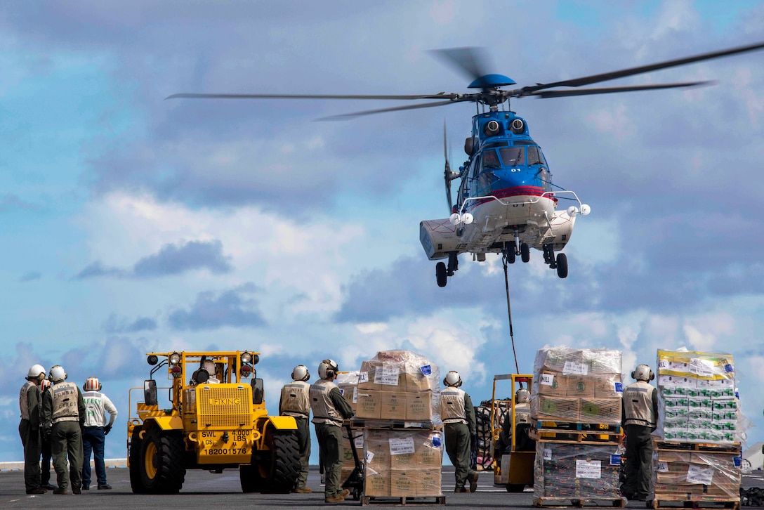 A helicopter hovers over the deck of a ship while sailors stand  around pallets.