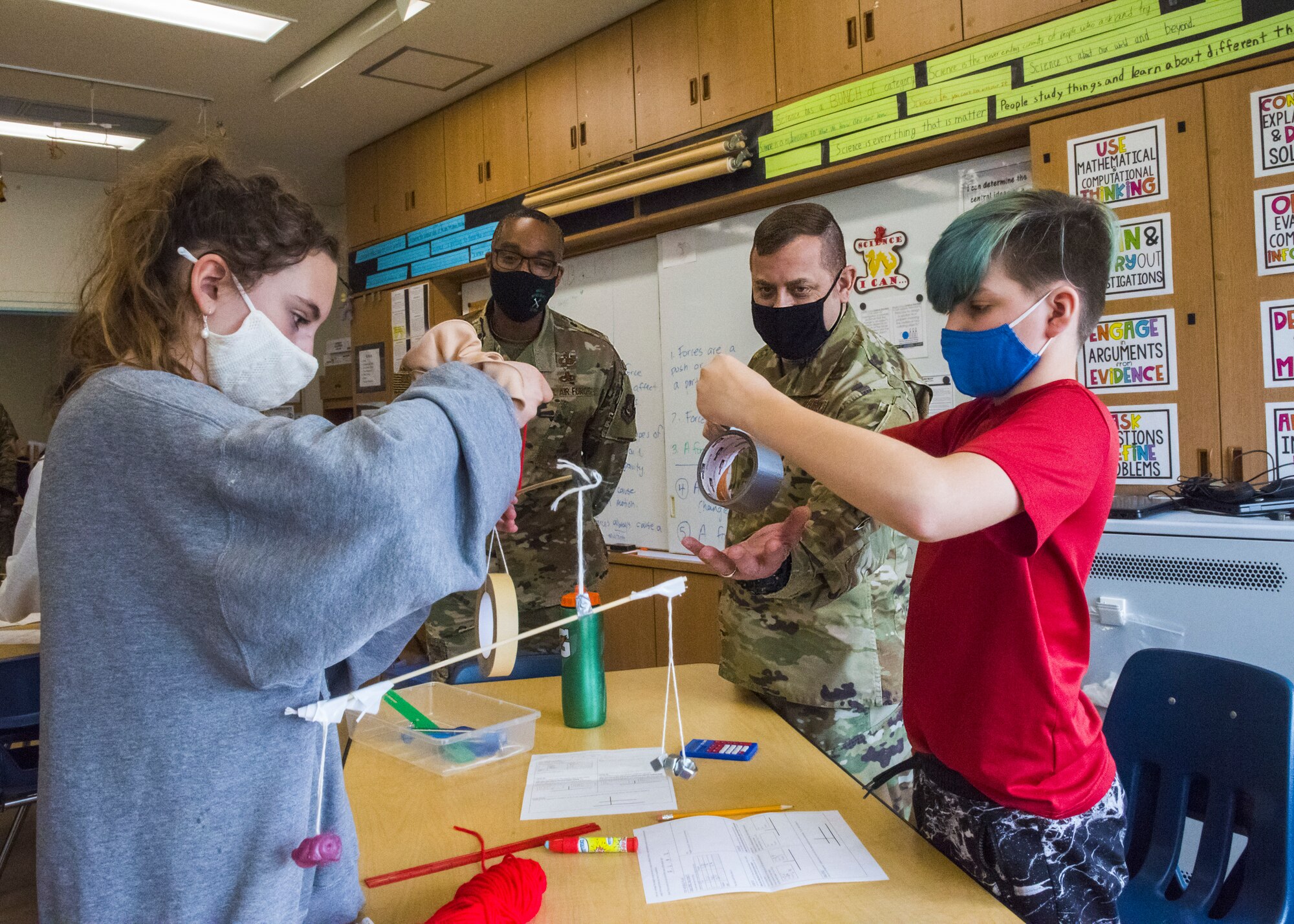 Two men in uniform assist two students, build a balancing contraption made from school supplies.