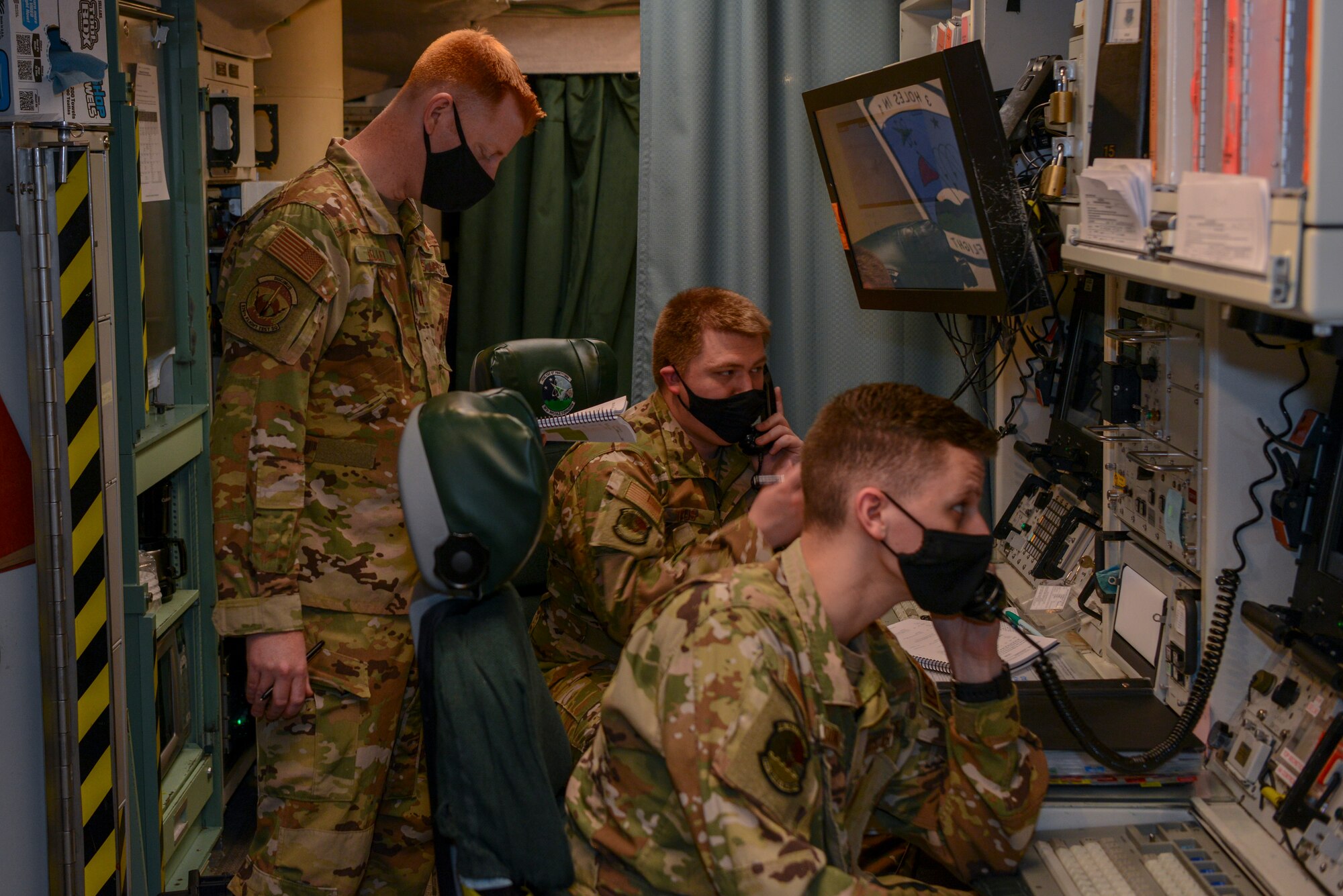 Two Airmen work at the launch console inside of a launch control center while another Airman observes