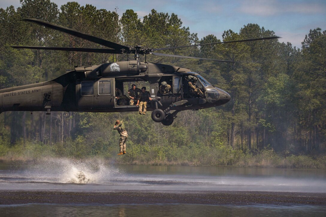 A soldier jumps out of a helicopter into a pond as fellow soldiers watch.