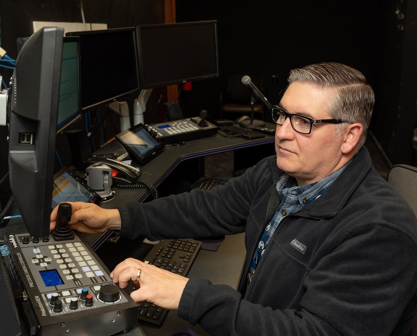 Photo of Grant Chyko sits at a computer console to set up one of the depots conference rooms for an upcoming meeting.  He's wearing black-framed glasses and a blue plaid shirt beneath a blue fleece jacket.