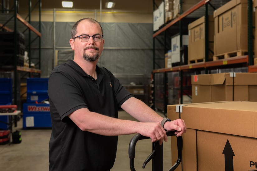 Photo of Andy Hrosovsky uses a hand truck to move equipment around a warehouse.  He is wearing black-framed glasses and a black short-sleeved polo shirt.