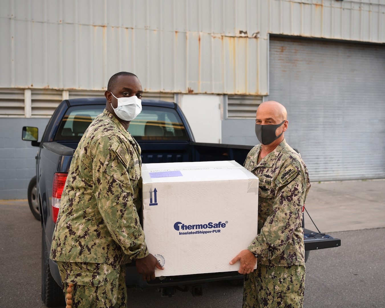 A white man and black man in camo uniforms hold a white box in front of a blue pickup truck.