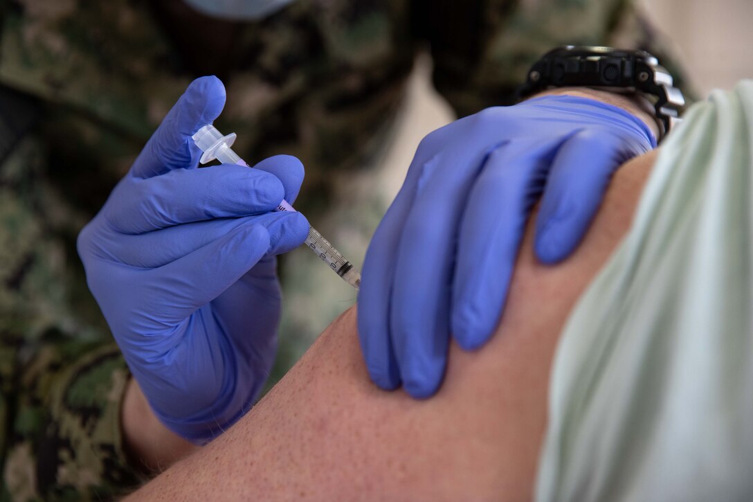 Two hands in blue gloves - one holds an arm while the other inserts a needle into the arm.