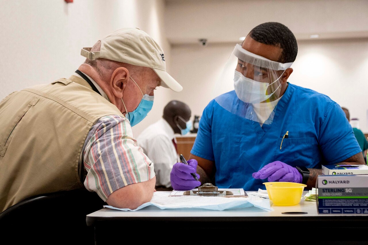 A sailor writes on a clipboard while sitting at a table next to another man.