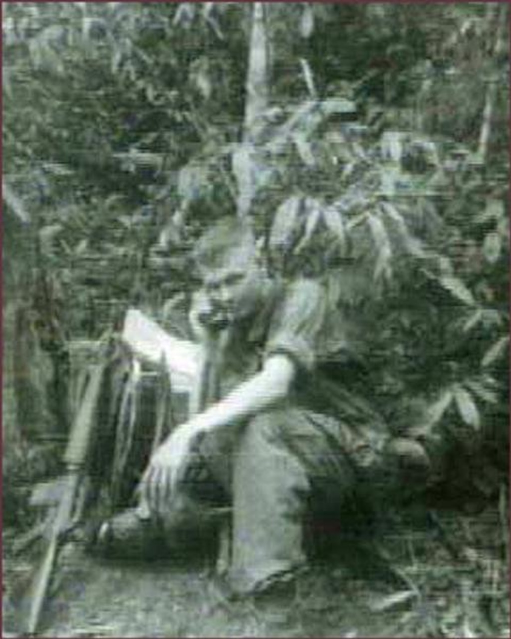 A teenager squats near a bush while holding a telephone to his ear. A rifle is propped near him.