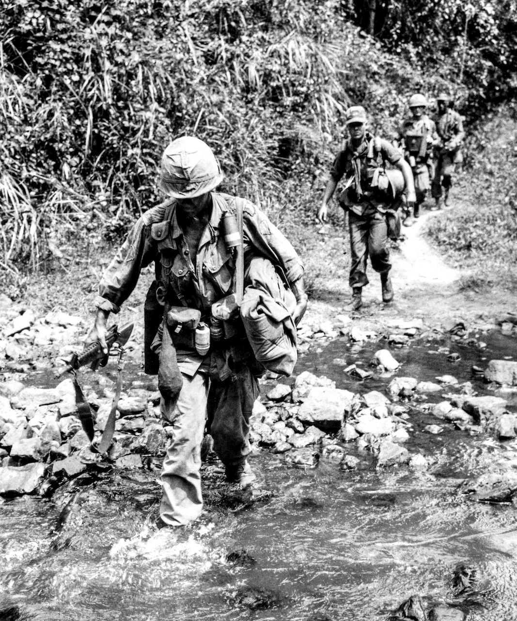 A man in combat uniform crosses an ankle-deep creek. Other men follow behind him on a trail coming out of the woods.