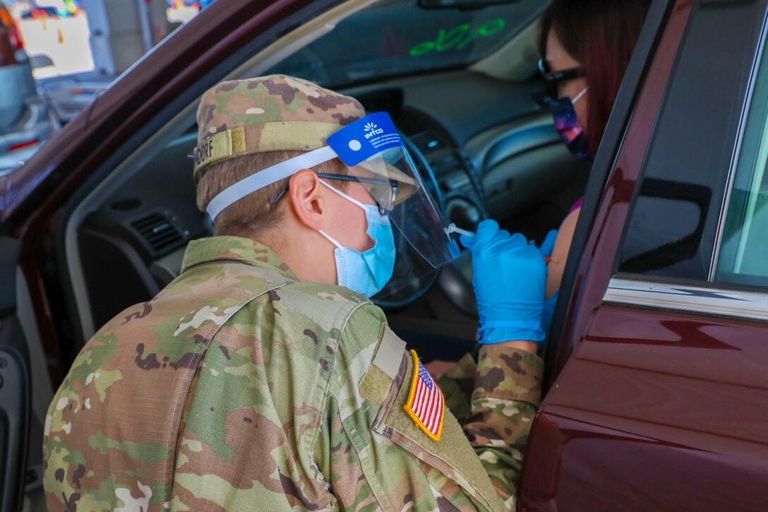 A person in military uniform kneels beside the driver's side of a passenger vehicle. She administers a vaccine to the driver.