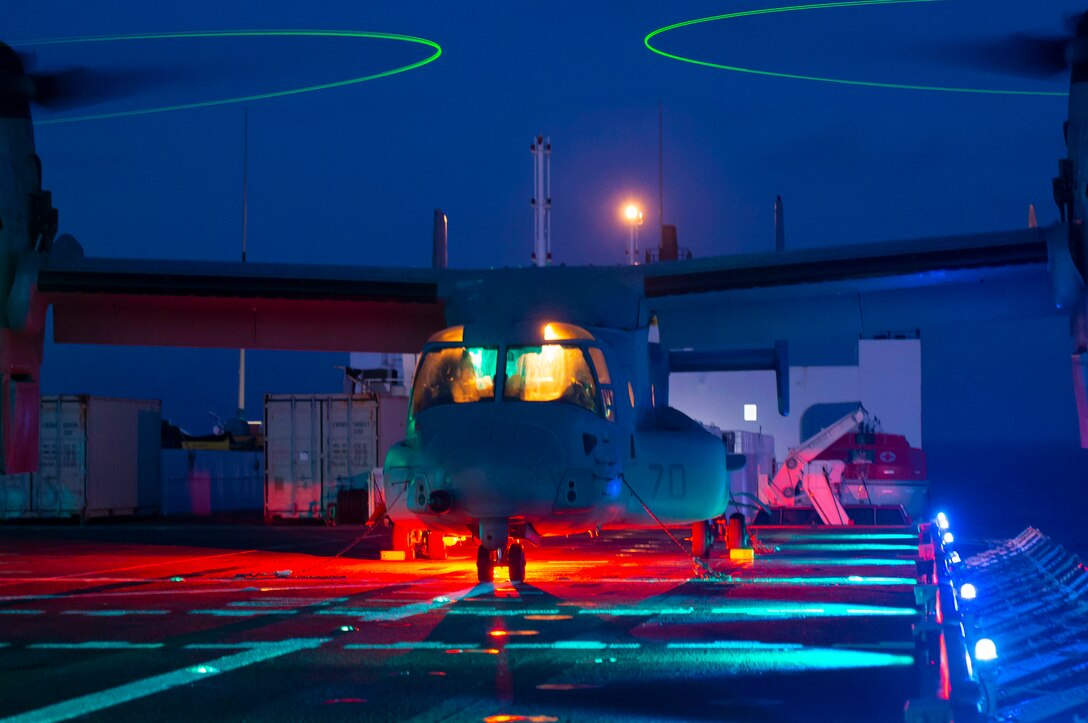 An aircraft surrounded by blue and red lights sits on the deck of a ship.