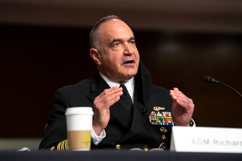 A man speaks at a hearing. A cup and microphone are in the foreground.