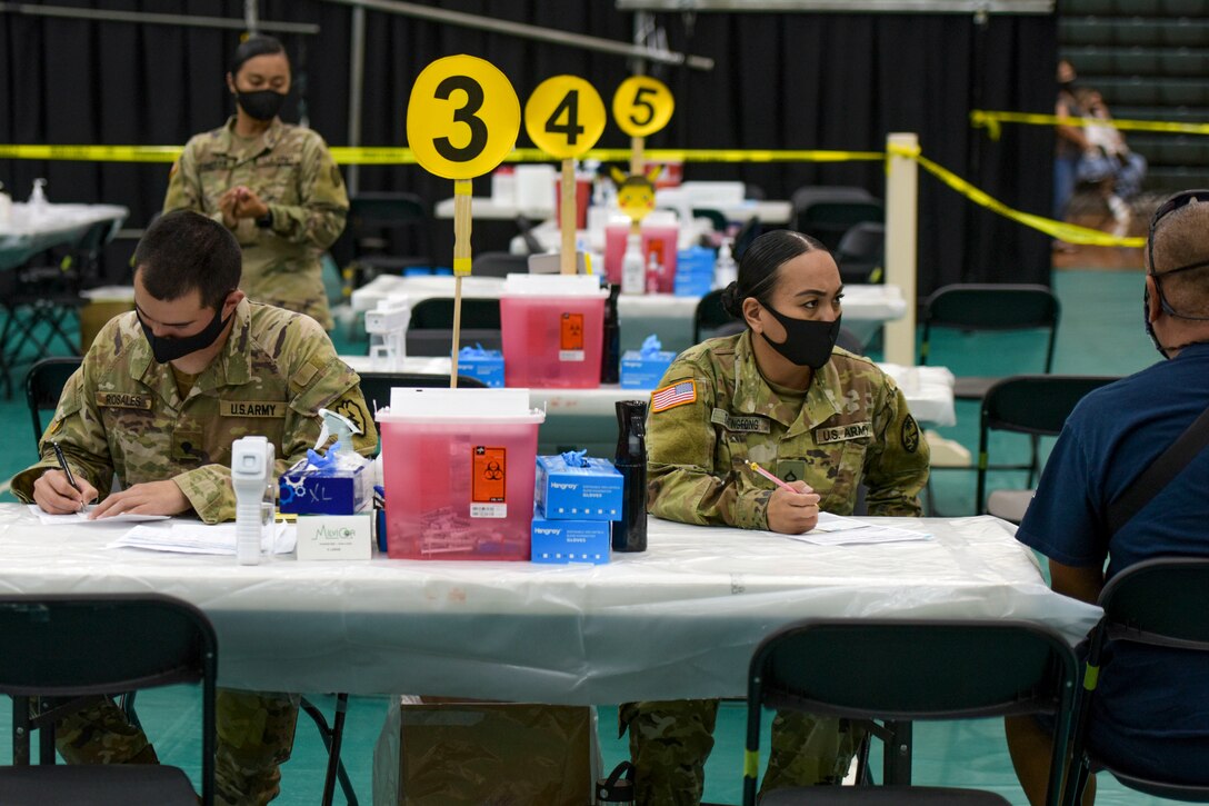 Two soldiers sit at a table, one talking to a patient, in a large room with other tables.