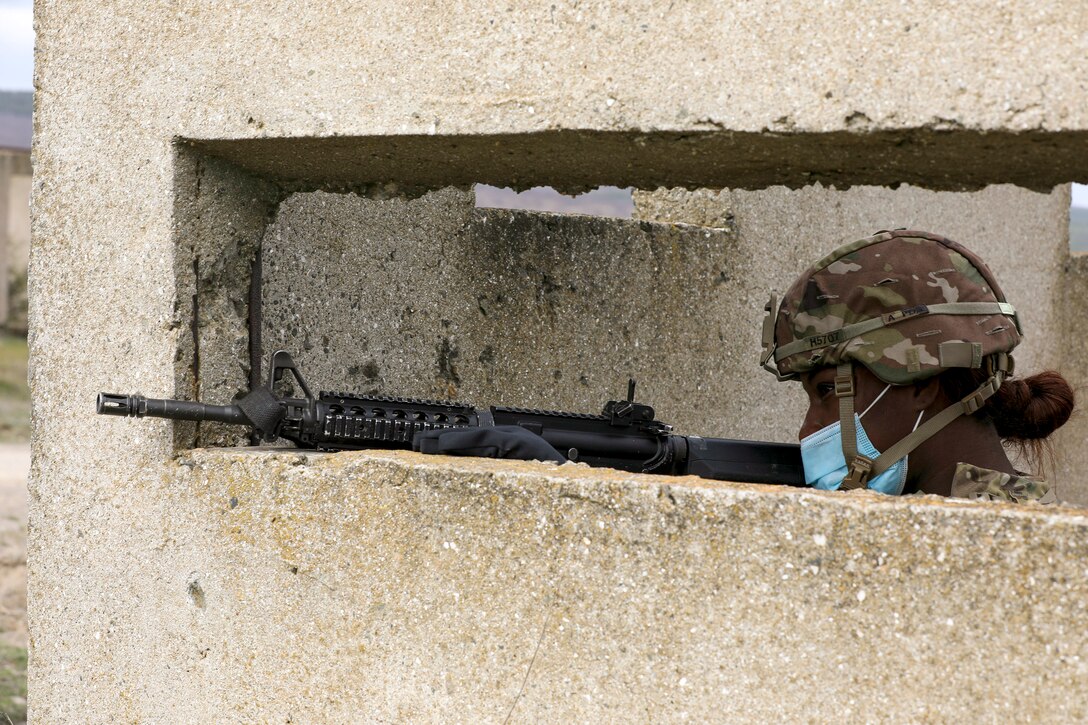 A soldier points a weapon and looks out from an opening in a concrete structure.