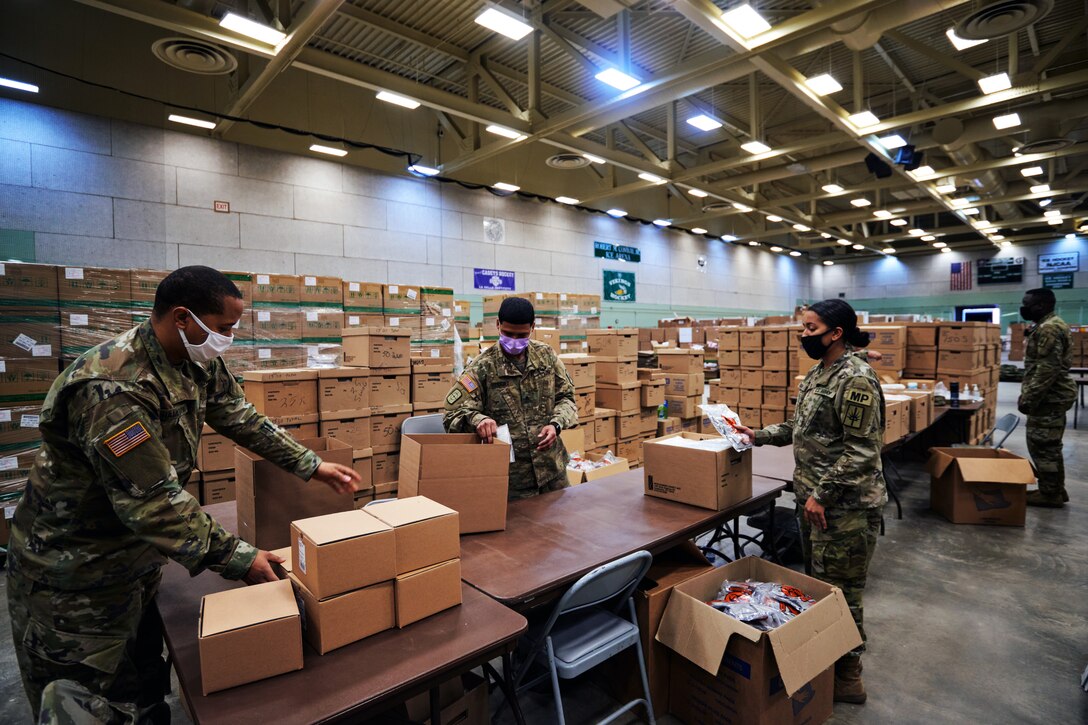 Three soldiers wearing face masks work at long tables in a room filled with large boxes; smaller boxes are on the table.