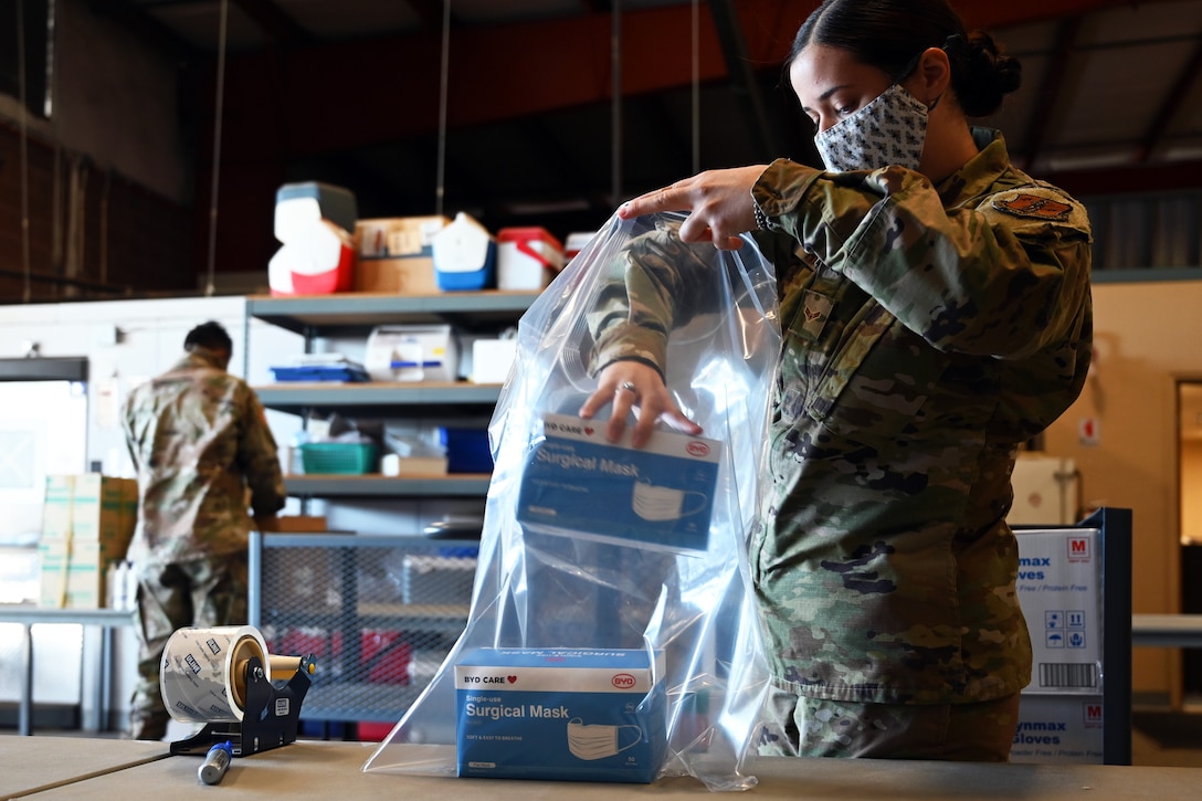 An airman wearing a face mask puts boxes that are labeled surgical masks into a large plastic bag.