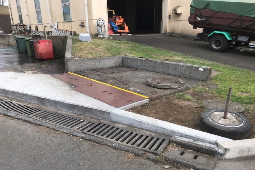 A curb and grating are seen outside a building; a vehicle and large trash cans are in the background.