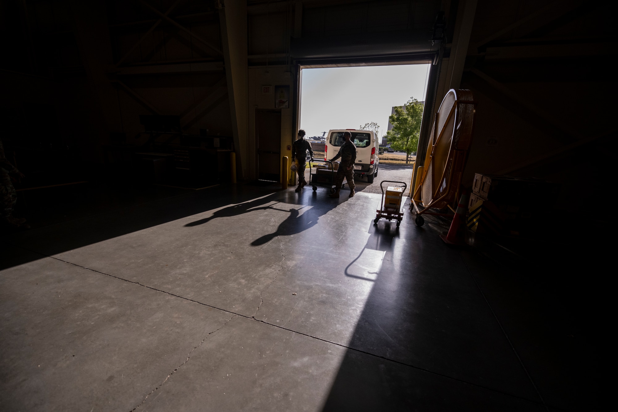 Two silhouettes of Airmen are seen walking in to a room from outside. Their shadows are seen on the floor. They are pushing a large cart covered with aircraft parts that are in need of repair.