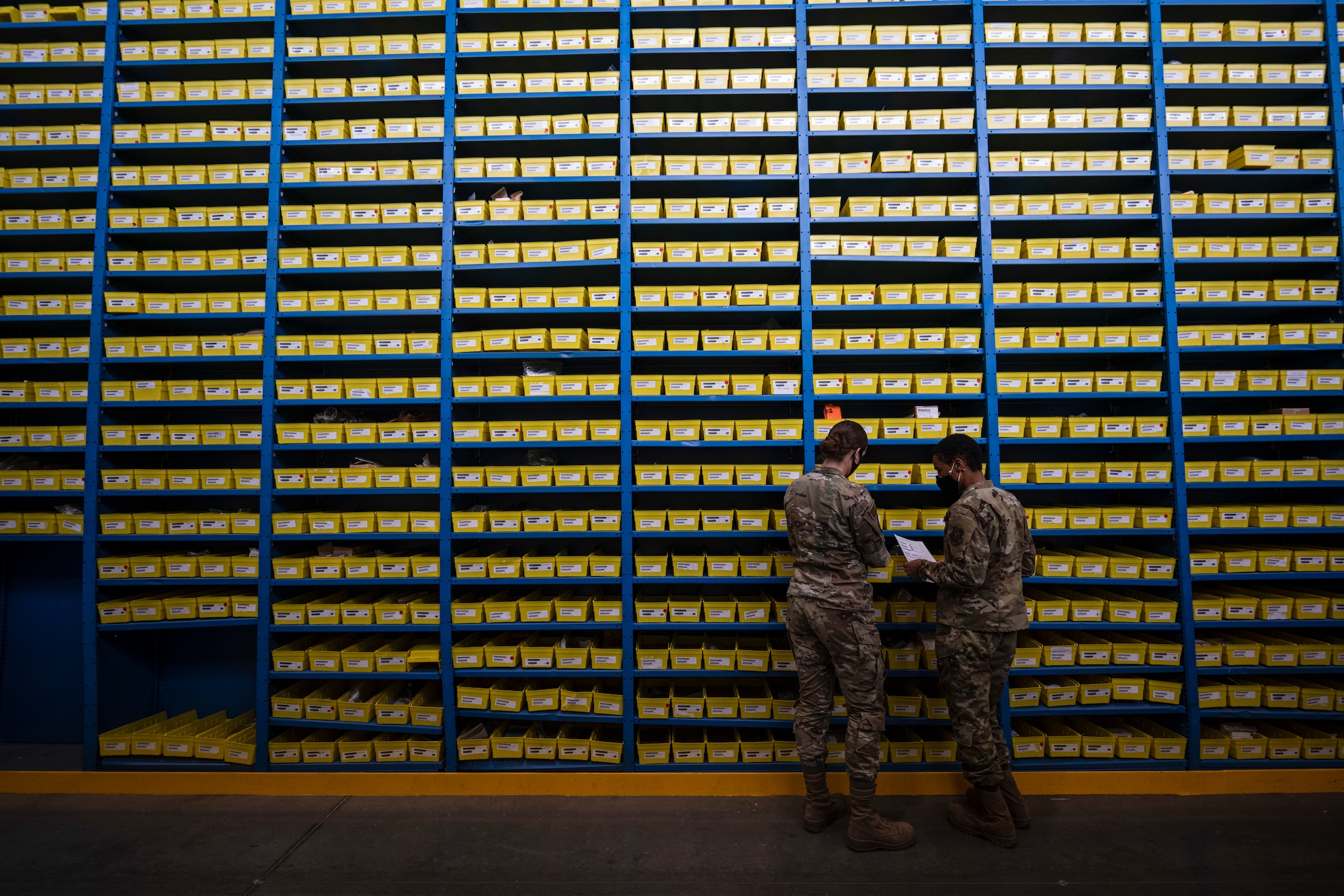 Two female Airmen stand next to each other next to a very tall wall of shelves
