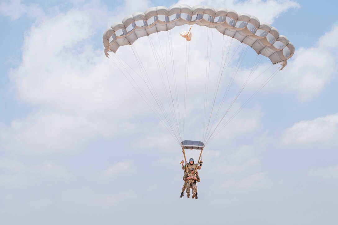 Two airmen wearing a parachute descend in the sky.