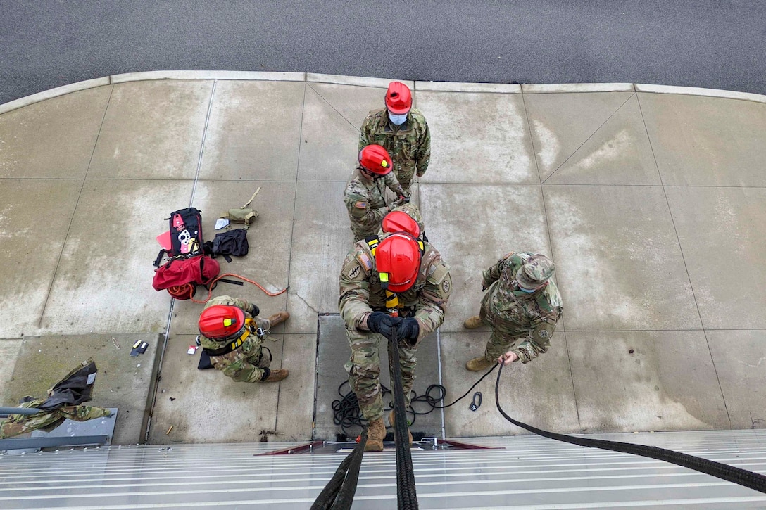 A guardsman holds onto a rope while climbing up the side of a buildings; others stand below.