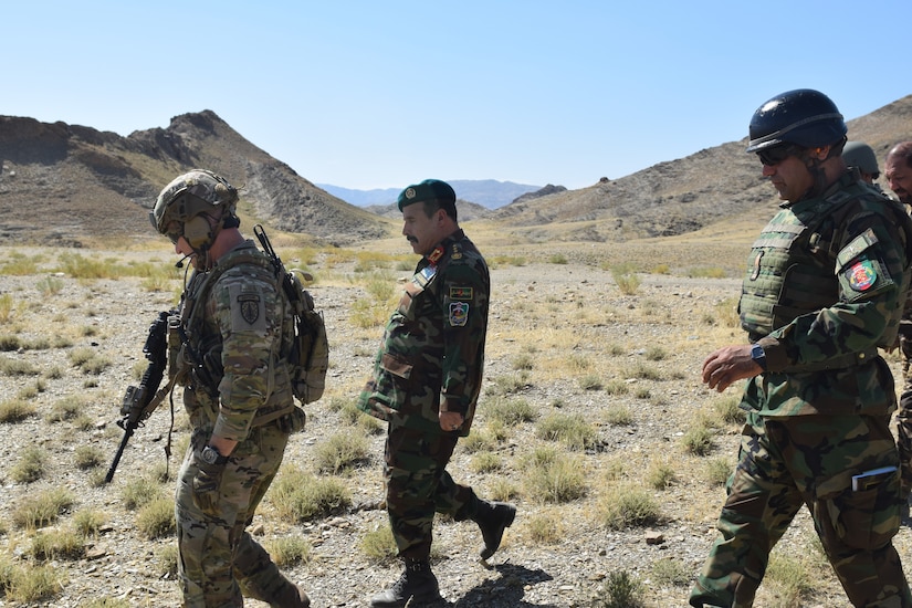 An American soldier and Afghanistan military personnel walk together across a rugged terrain.